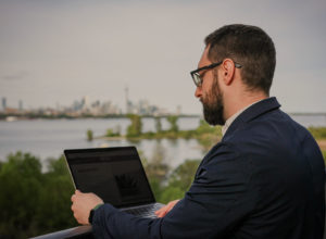 Man with laptop working outside. Big city in background across a lake.